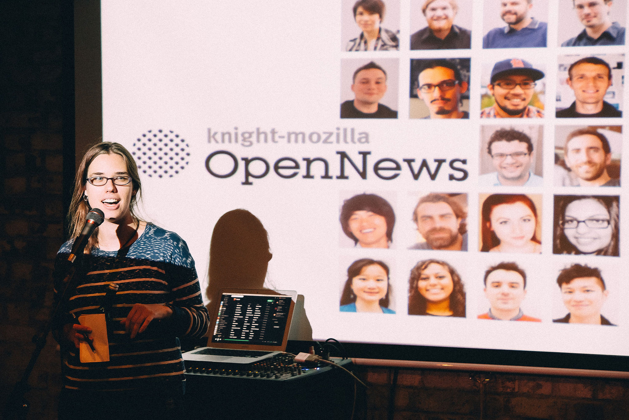 Erika (a bespectacled white person with messy hair) is standing in a striped jumper addressing a crwod in front of an image array of the Knight-Mozilla Fellows.