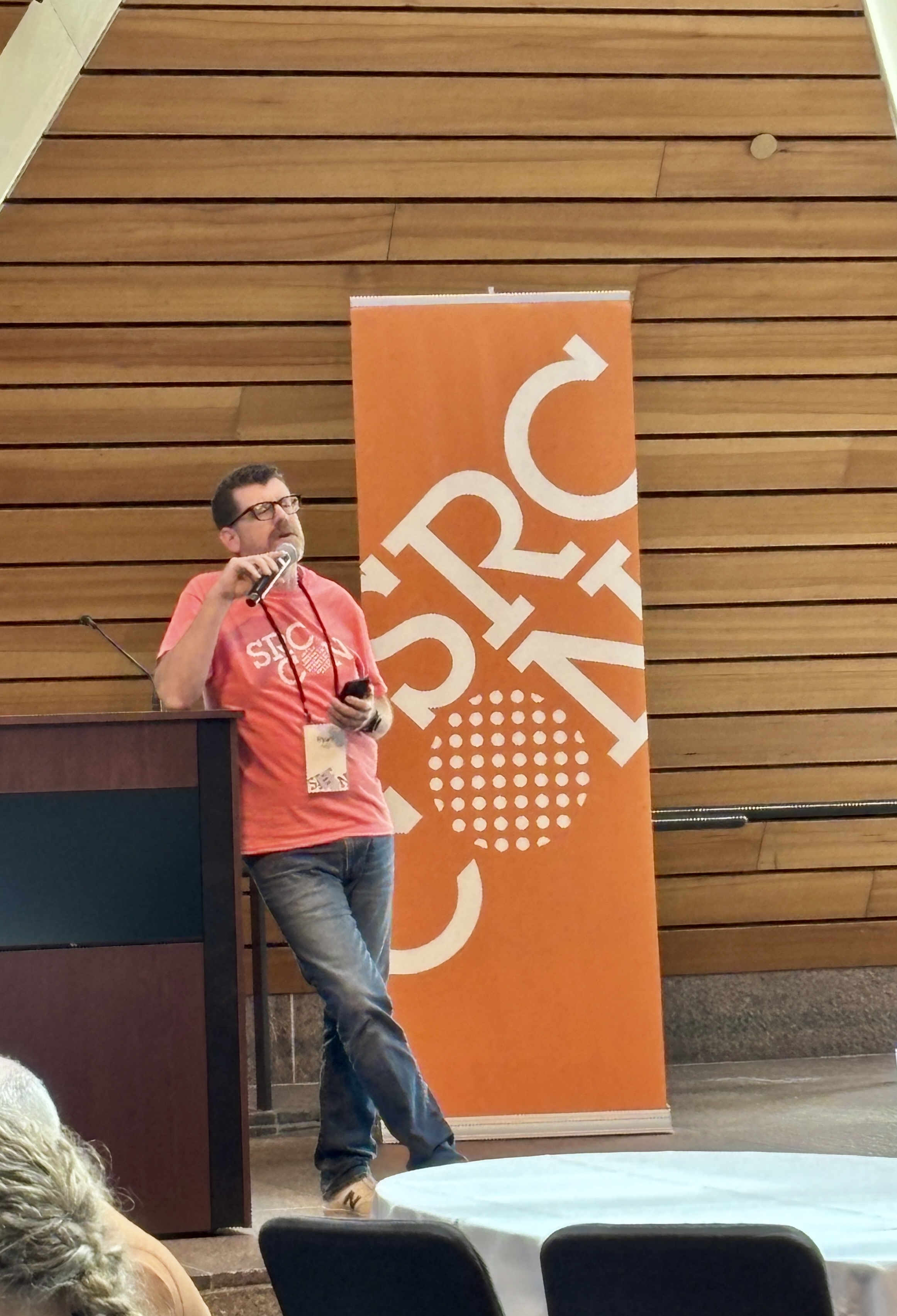 Ryan (a bespectacled white person with a beard) is leaning against a podium, holding a microphone in front of an orange sign that says SRCCON