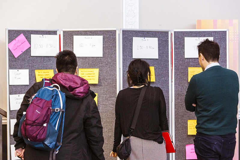 three people with their backs to the camera in front of a bunch of big post its organized as a conference schedule